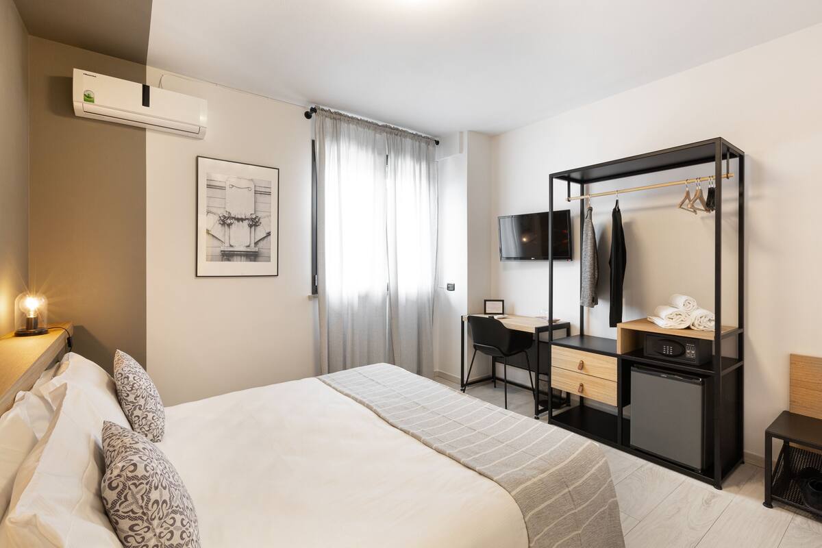 A hotel room with a bed, desk, chair, TV, wardrobe, and a small fridge.