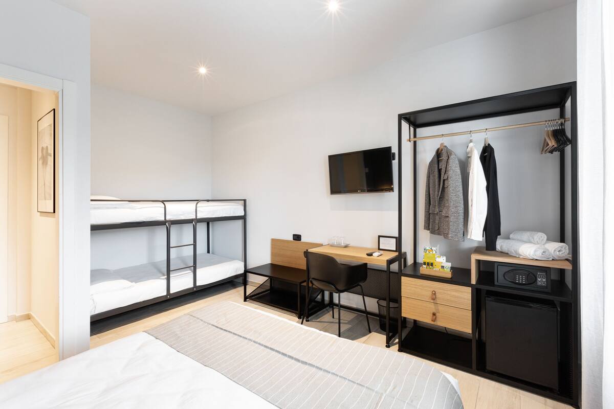 A modern hotel room with bunk beds, a desk, a chair, a TV, a wardrobe, and a mini-fridge.