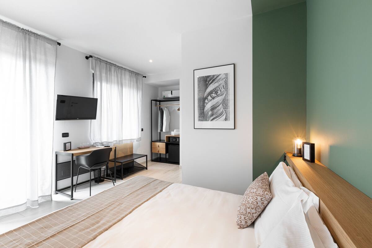 A modern hotel room with a bed, desk, and chair, a flat-screen TV, and a framed artwork on the wall.