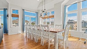 A spacious dining area with a wooden table and white chairs, large windows with blue curtains, and a view of neighboring houses.