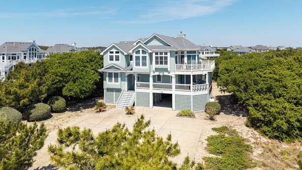 A large, multi-story house with a balcony, surrounded by dense greenery and a sandy driveway.