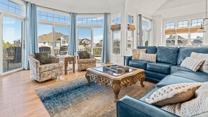 A spacious living room with a large blue sofa, two patterned armchairs, a wooden coffee table, and a view of neighboring houses through large windows.