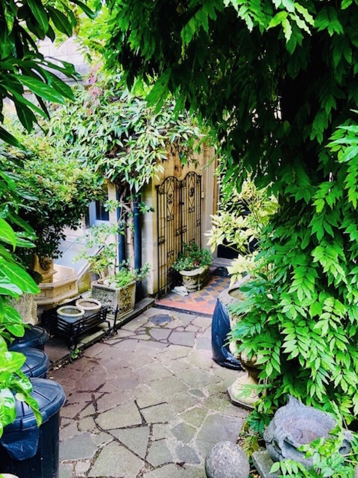 A stone-paved courtyard with a wrought iron gate, surrounded by lush greenery and potted plants.