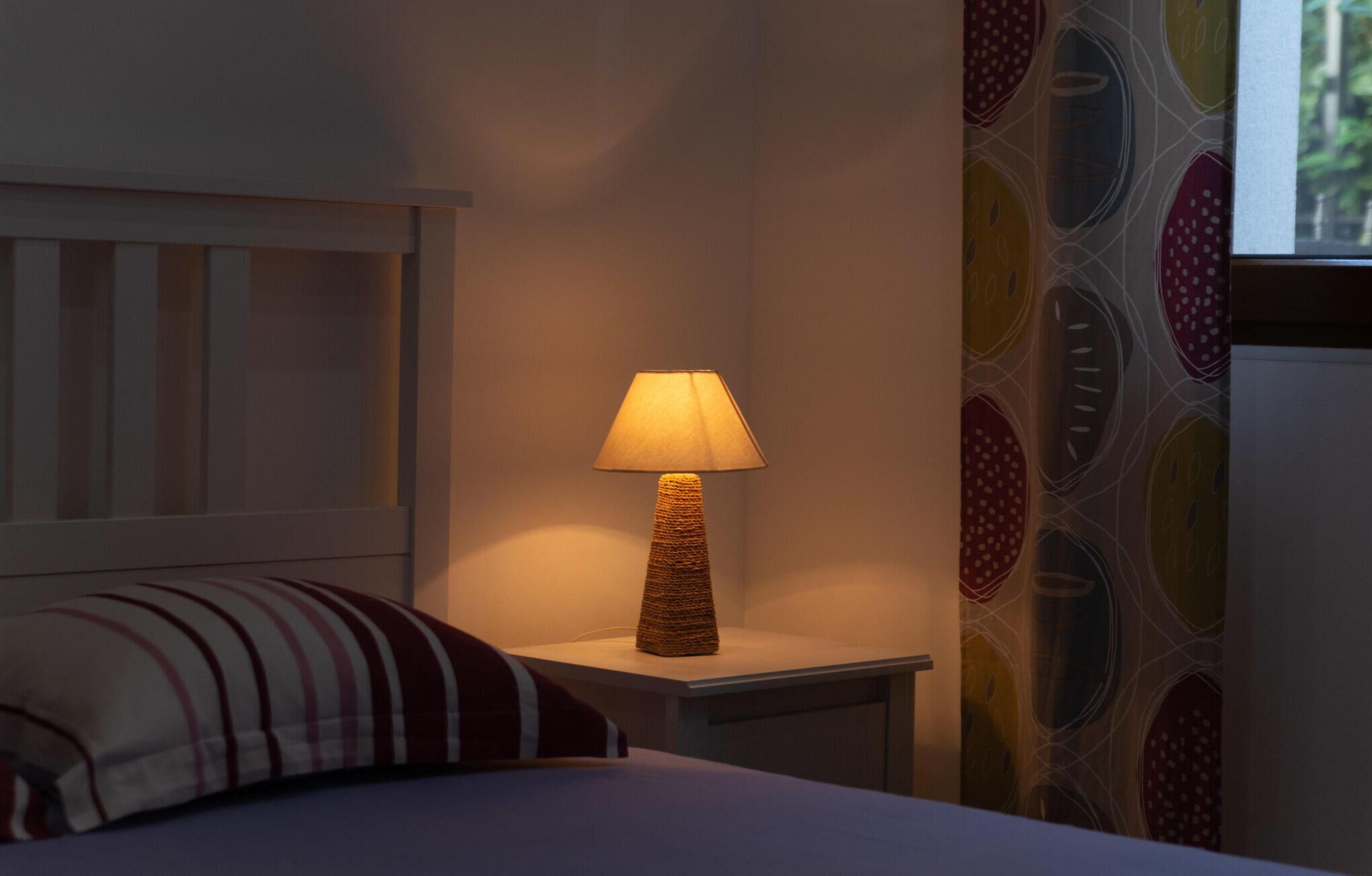 Un lit avec un oreiller à rayures, une table de chevet surmontée d’une lampe en osier, et un mur orné d’un motif circulaire aux couleurs vives.