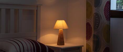 Un lit avec un oreiller à rayures, une table de chevet surmontée d’une lampe en osier, et un mur orné d’un motif circulaire aux couleurs vives.
