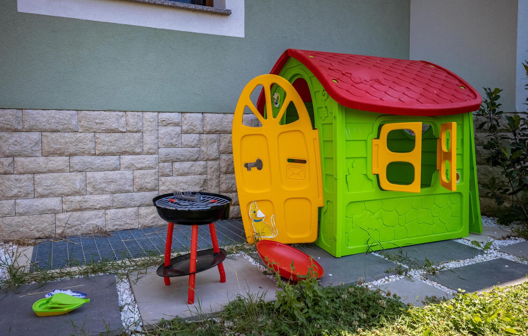 Una casita de juegos infantil con techo rojo, paredes verdes, una parrilla negra pequeña y una puerta amarilla.
