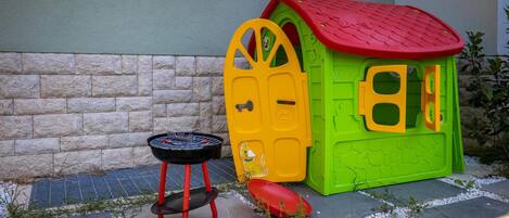 Una casita de juegos infantil con techo rojo, paredes verdes, una parrilla negra pequeña y una puerta amarilla.
