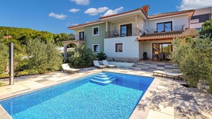 Uma casa de dois andares com piscina, área de estar externa e vista para o verde ao redor.