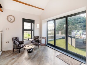 A spacious living room with a large window, a round coffee table, two leather armchairs, and a wall clock.