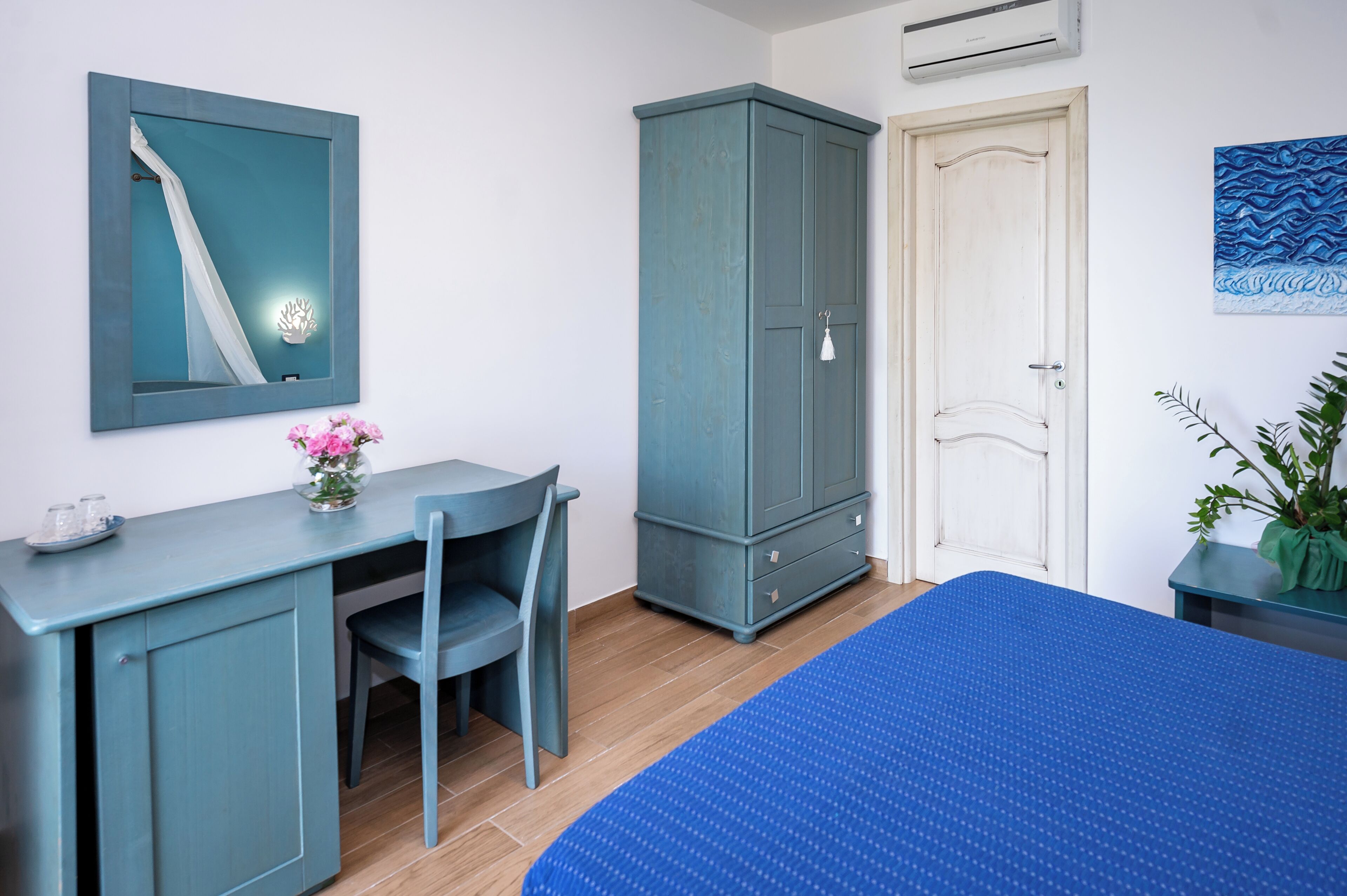 Een kamer met een blauw bed, een bureau met stoel, een kledingkast, een deur en een wandairconditioner.