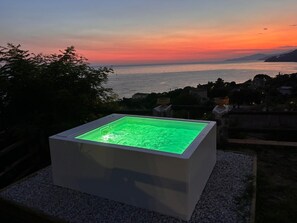 A square hot tub with a green light, set on a gravel surface, overlooking a body of water and a sunset.