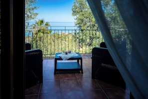 A balcony with wicker furniture, a small table with a lace tablecloth, and a view of the sea and greenery.