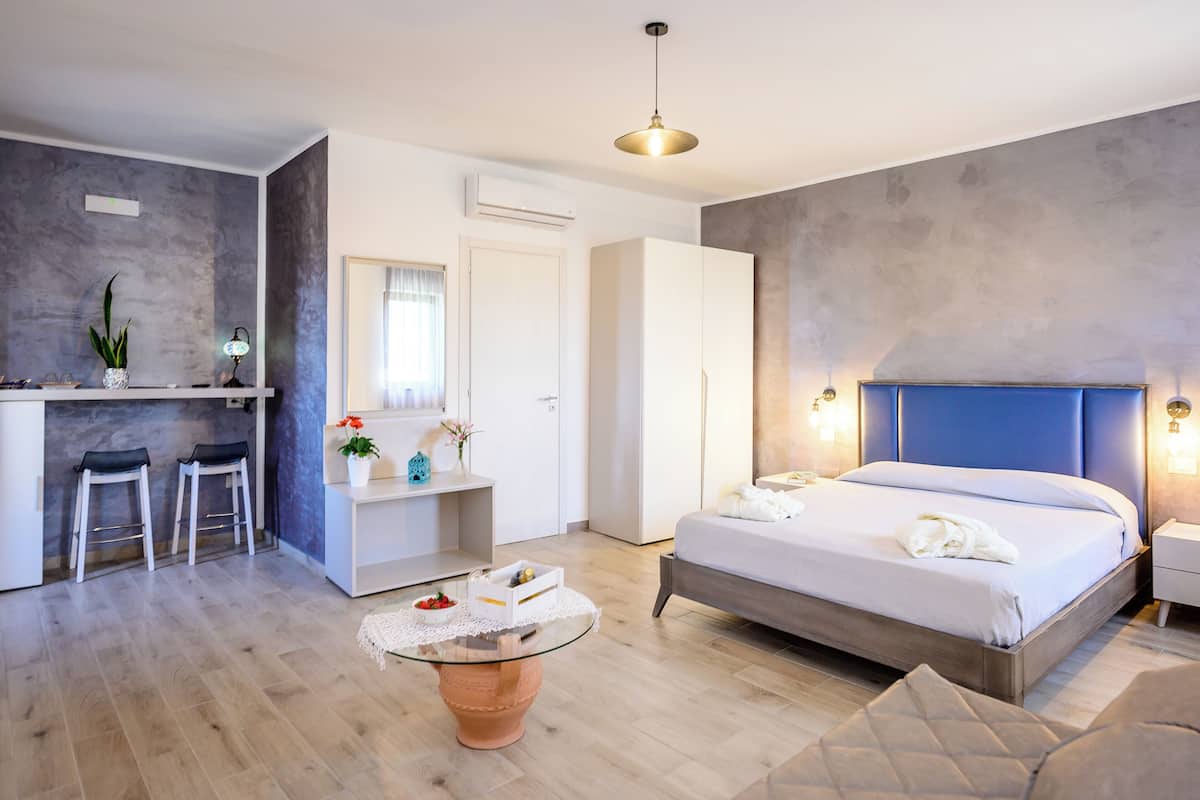 Moderni hotellihuone, jossa on sänky, yöpöydät, pieni työpöytä, tuoli ja ruukkukasvi.