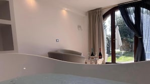 A bedroom with a large bed, a chair, a small table with bottles, and a view of greenery through a window.