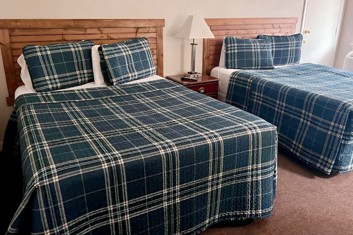 A hotel room with two beds, each with matching plaid bedding, a wooden headboard, a bedside table with a lamp, and a door with a golden handle.