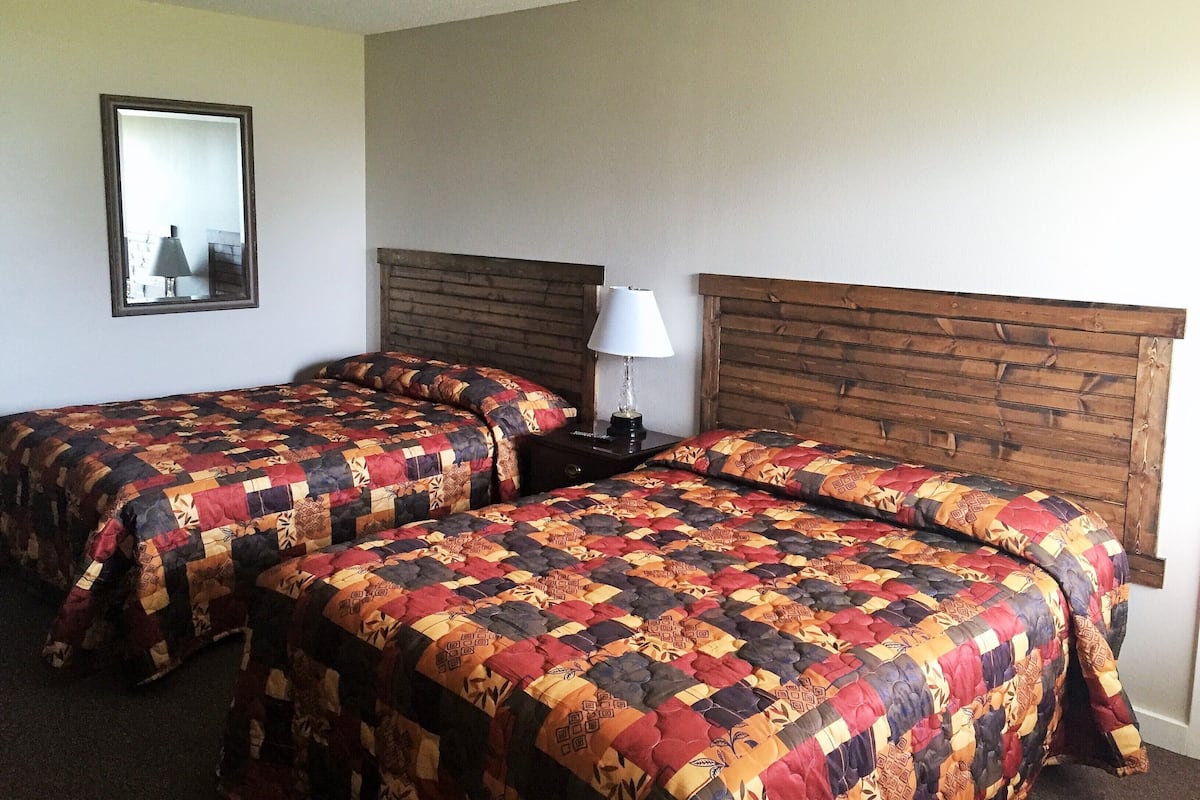 A hotel room with two beds, each with a colorful quilt, a wooden headboard, a nightstand with a lamp, and a mirror on the wall.