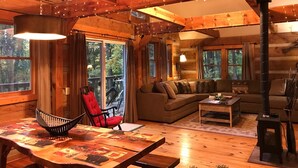 A cozy cabin interior with a wooden dining table, a red chair, a sofa, and a fireplace.