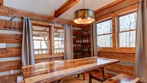 A rustic wooden interior with a large wooden table and benches, large windows with curtains, and a hanging light fixture.