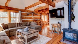 A cozy log cabin interior with a wooden ceiling, a large sofa, a coffee table, a fireplace, and a television mounted on the wall.