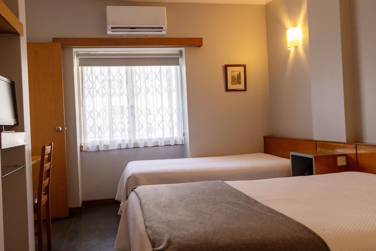 Habitación de hotel con dos camas, una ventana con cortina, aire acondicionado de pared y un cuadro enmarcado en la pared.