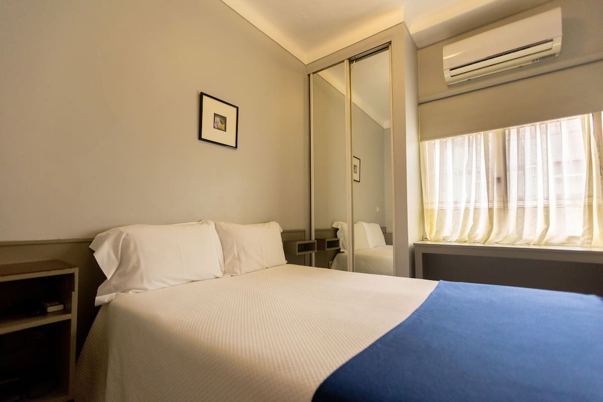 Una habitación de hotel con una cama, una mesita de noche, un espejo y un equipo de aire acondicionado.