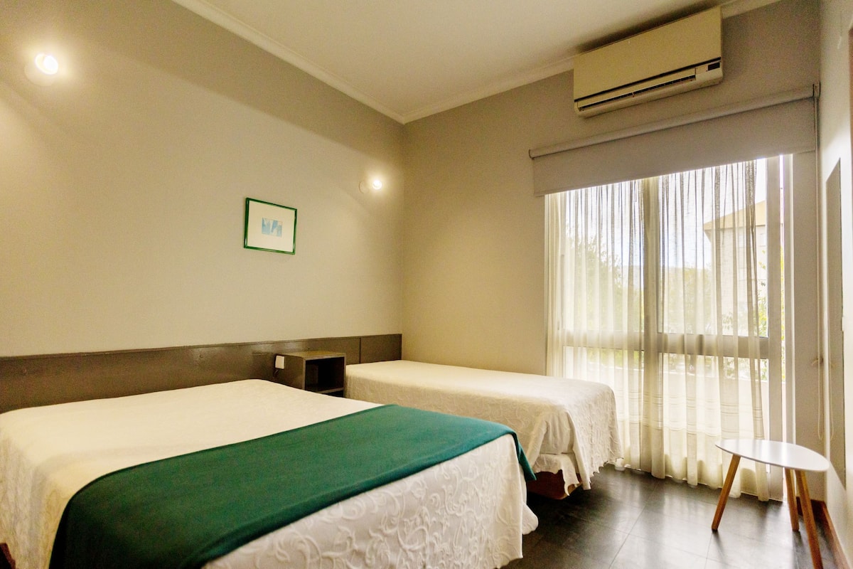Habitación de hotel con dos camas individuales, una mesita, una lámpara de noche y una ventana con cortinas traslúcidas.
