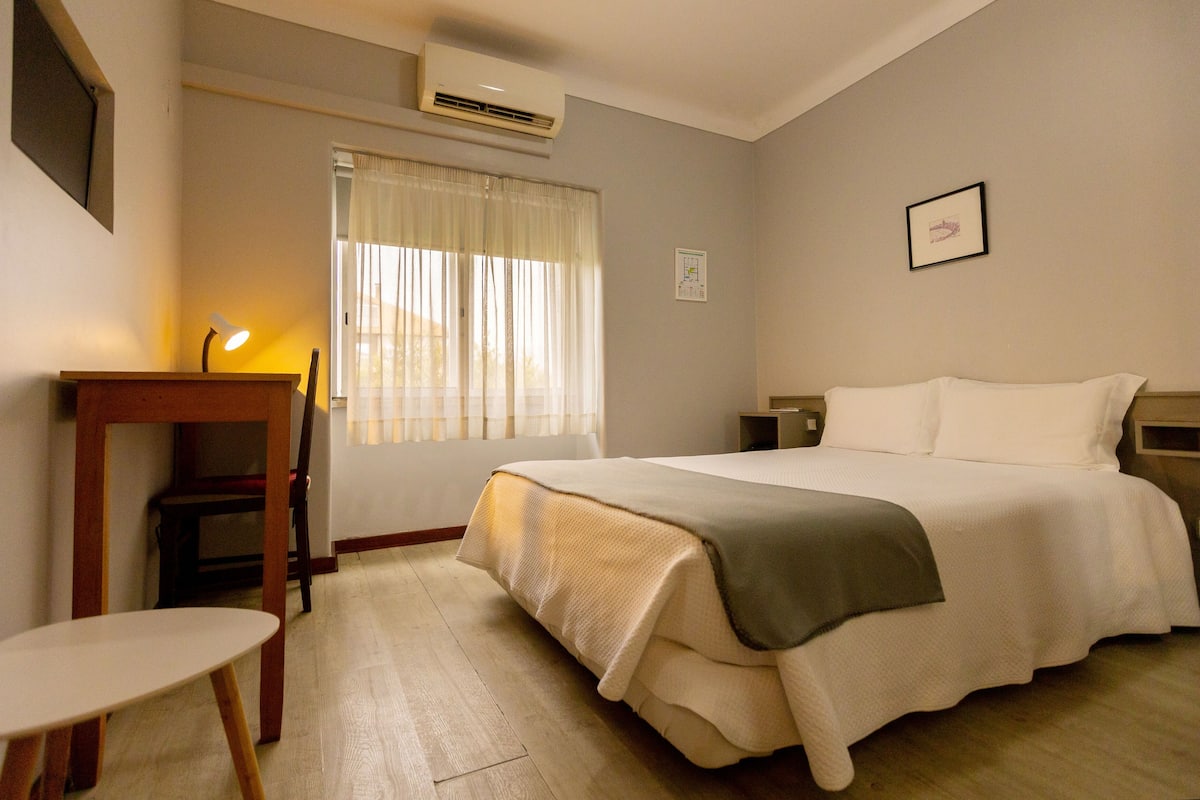 Habitación de hotel con cama, mesita de noche, lámpara y ventana con cortinas.