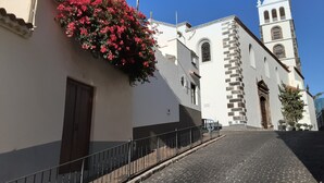 Eine Kopfsteinpflasterstraße führt zu einem weißen Gebäude mit einem Glockenturm, einem Blumenkasten mit rosa Blumen und einem Metallzaun.