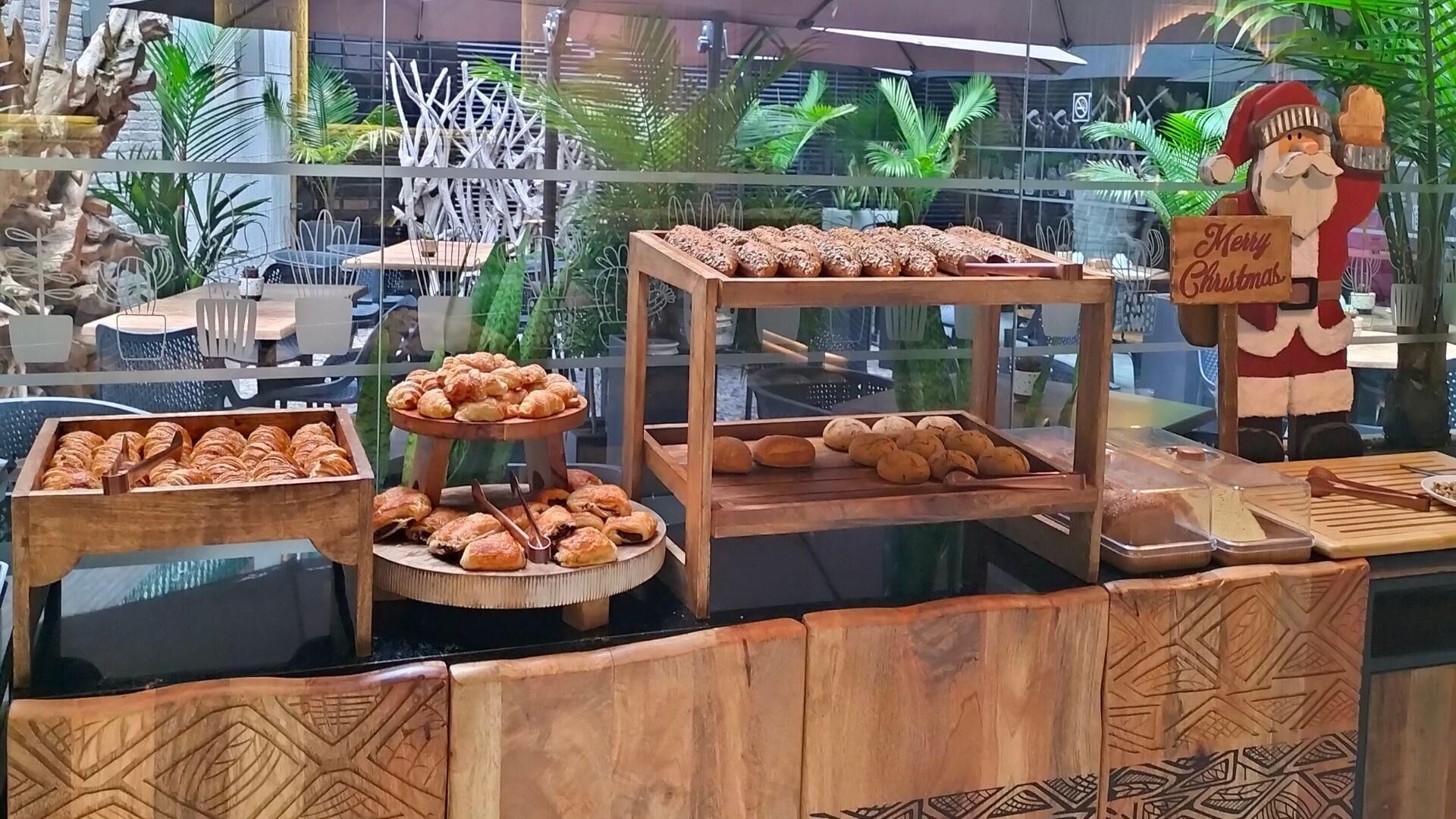 Una vitrina de panadería con diversos productos horneados, incluyendo croissants y pan, en un espacio con escaparate de vidrio, plantas decorativas y la figura de Papá Noel.
