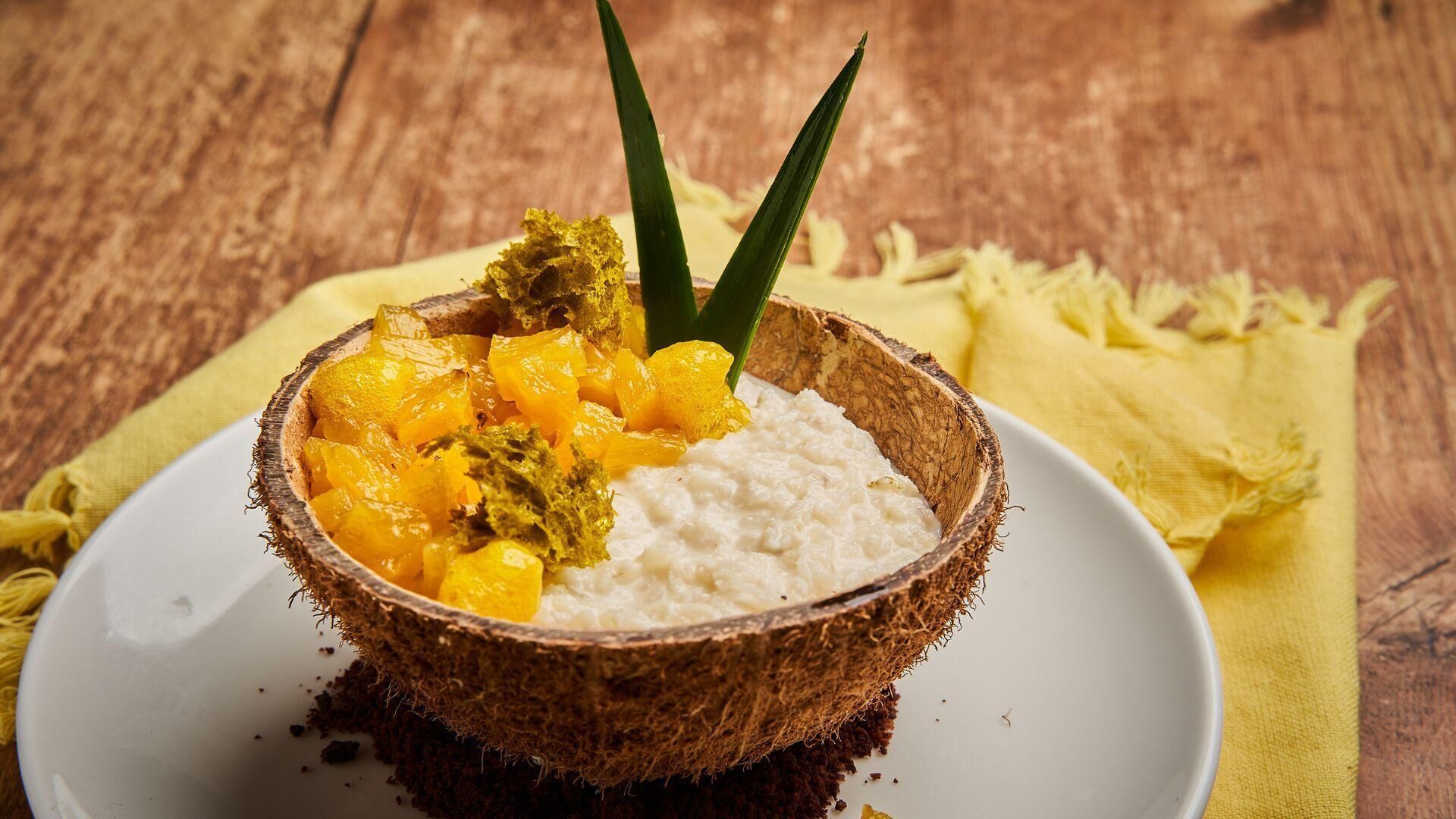 A bowl of coconut rice with mango chunks and green herbs on a white plate, placed on a wooden table with a yellow cloth.