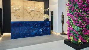 Un área de recepción de diseño moderno, con un gran panel de madera con relieves abstractos, una sección de mármol azul y una pared con flores artificiales coloridas.