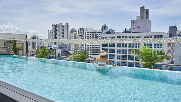 Una piscina en la azotea con agua azul cristalina, rodeada de palmeras y un horizonte urbano al fondo.