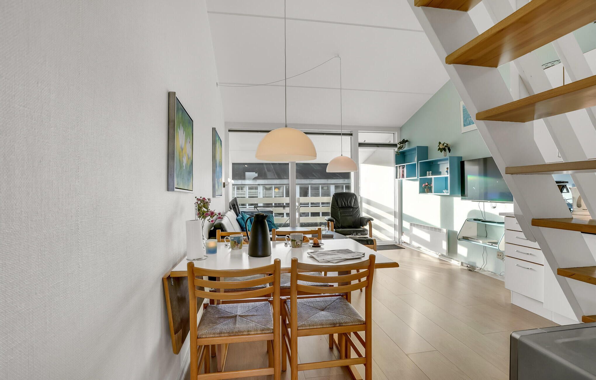 Et moderne spiseområde med et træbord og stole, et stort vindue med naturligt lys og en trappe med et geometrisk design.
