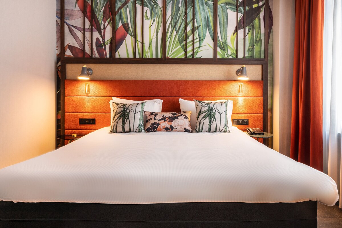 A hotel room with a large bed, two bedside tables, a wall-mounted lamp, and a decorative plant wall.