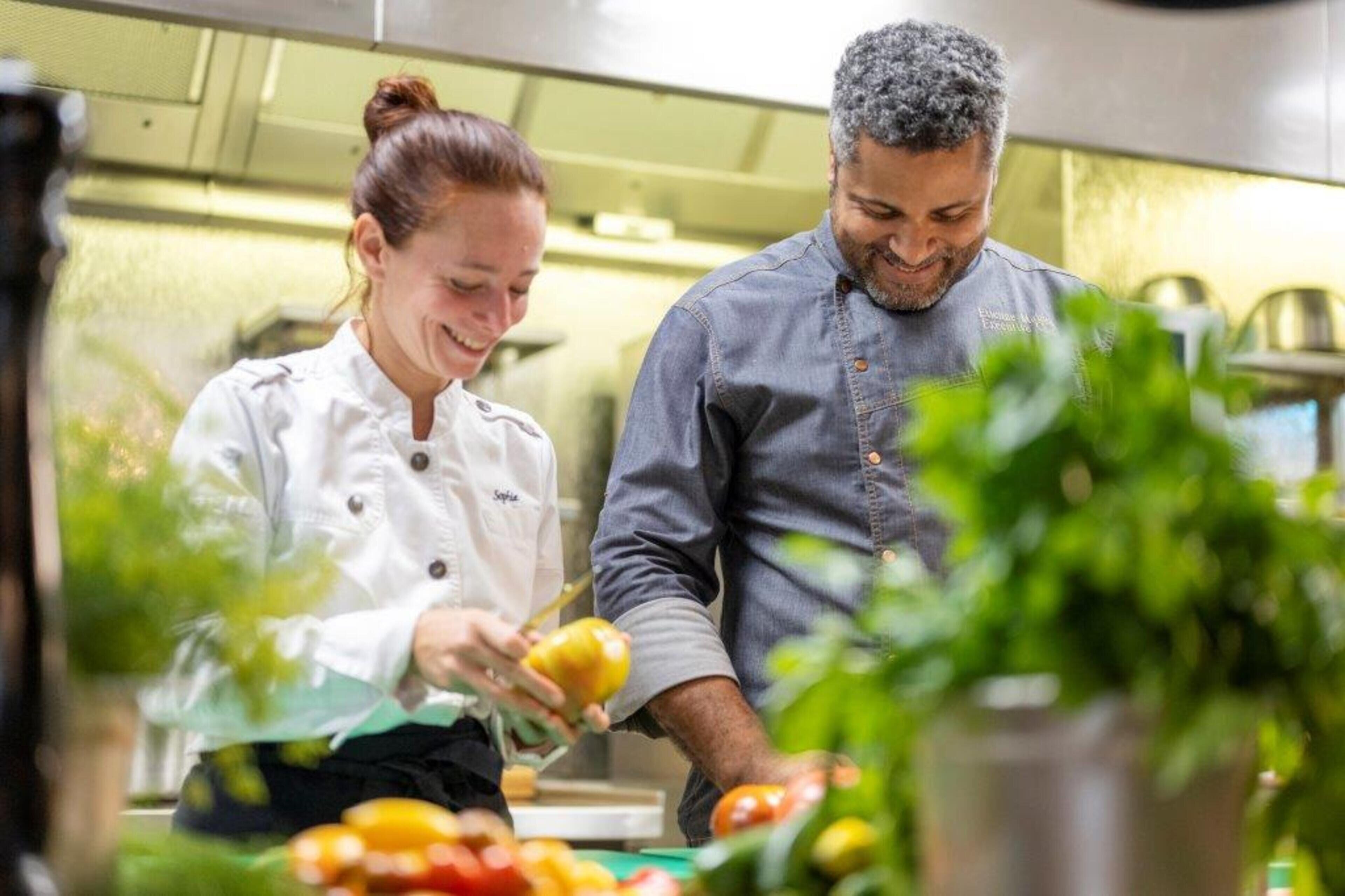 Due persone in cucina, una con un camice da chef e l'altra con abiti casual, stanno preparando del cibo.