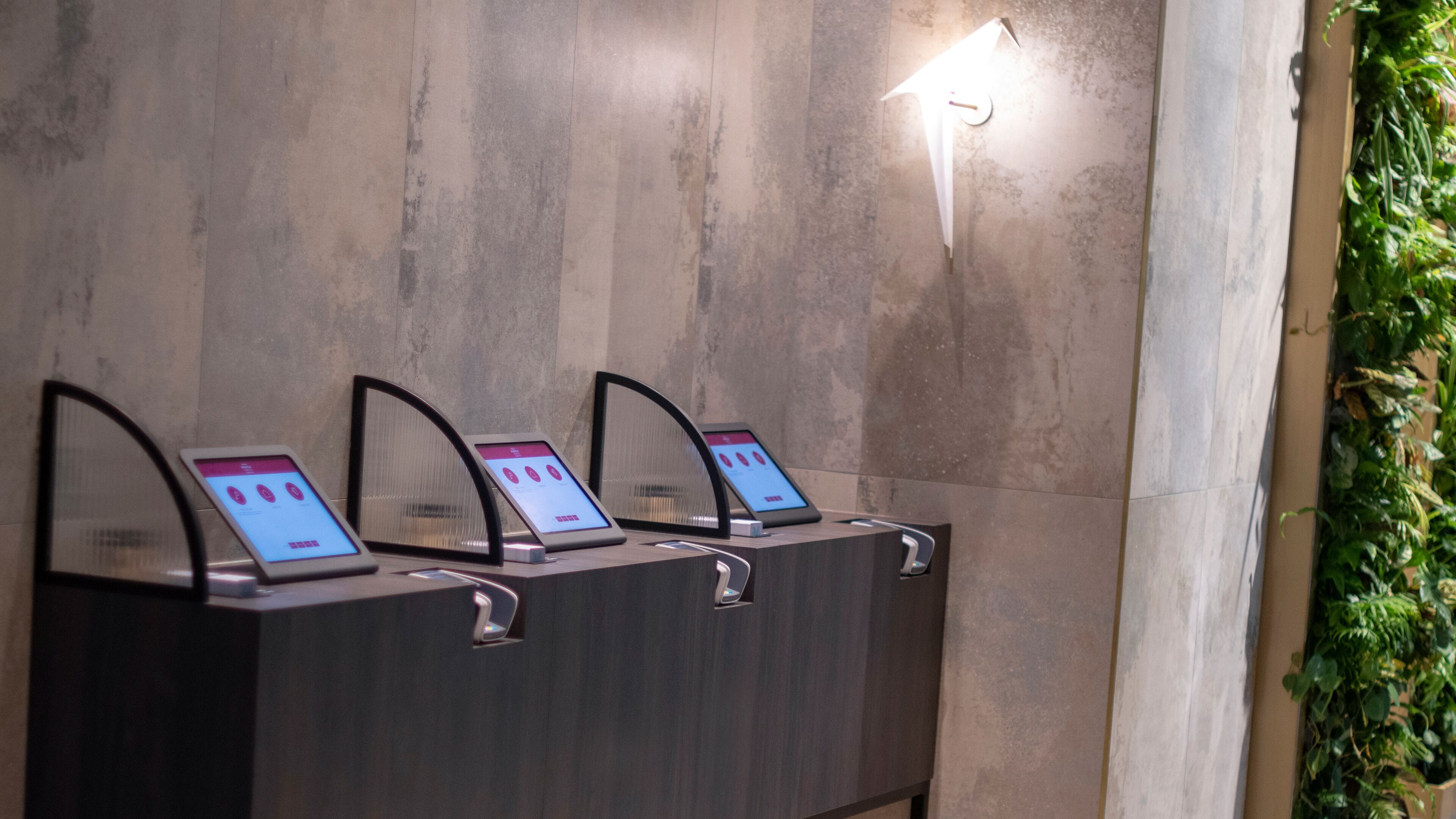 Een rij automaatkiosken met digitale schermen, voor een muur met structuur en een verticale lamp.