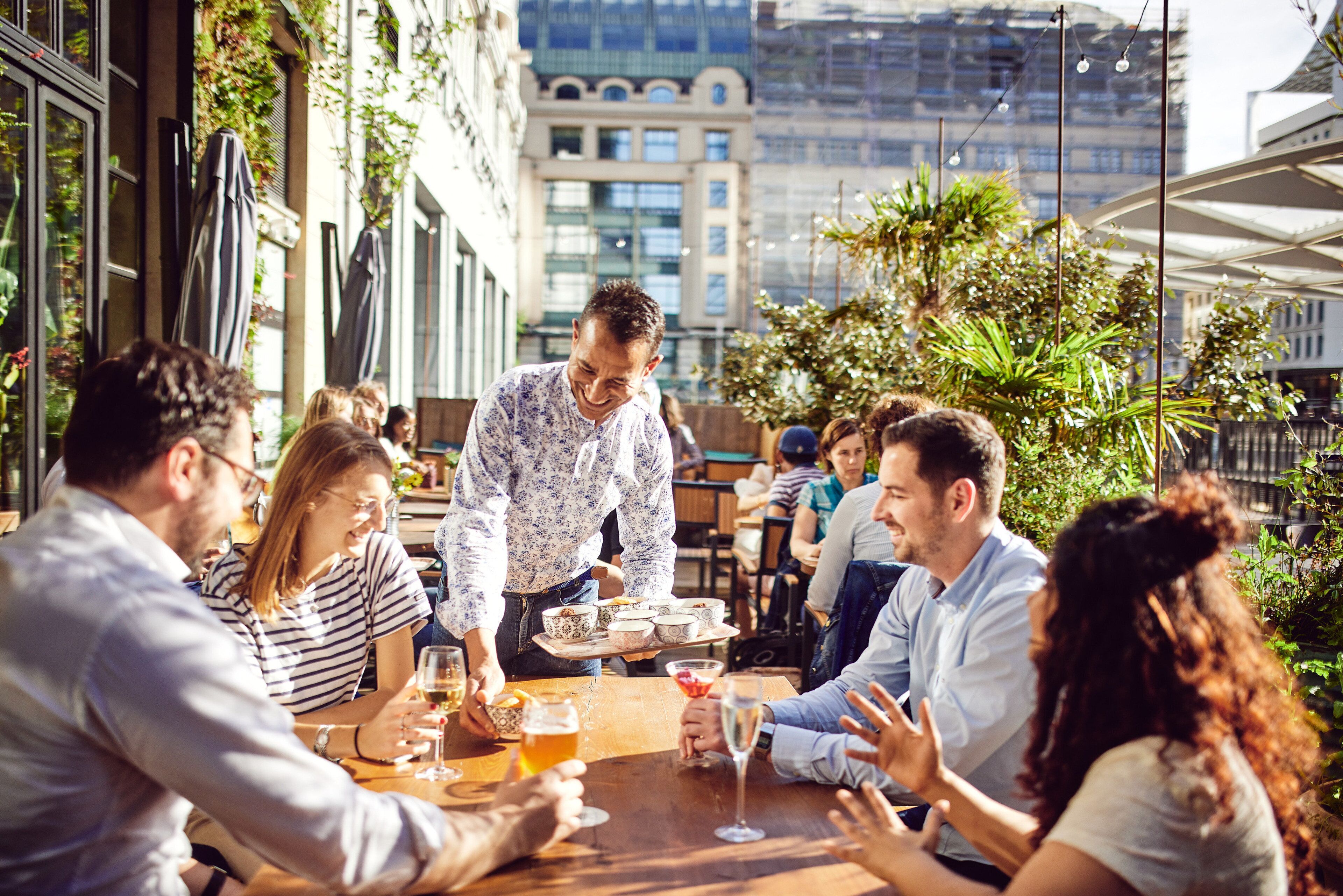 A group of people dining outdoors at a restaurant with a waiter serving drinks.