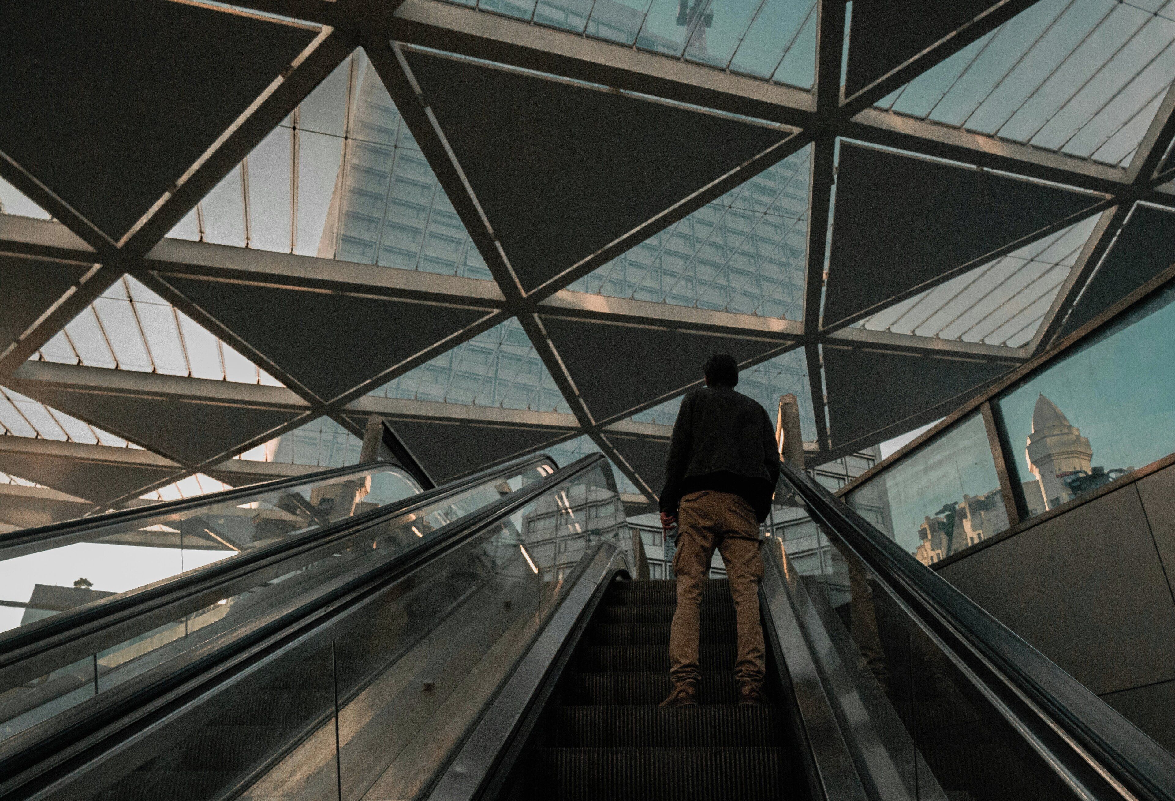 Une personne se tient sur un escalier mécanique à l’intérieur d’un bâtiment dont l’architecture moderne est caractérisée par l’utilisation du verre et de l’acier.