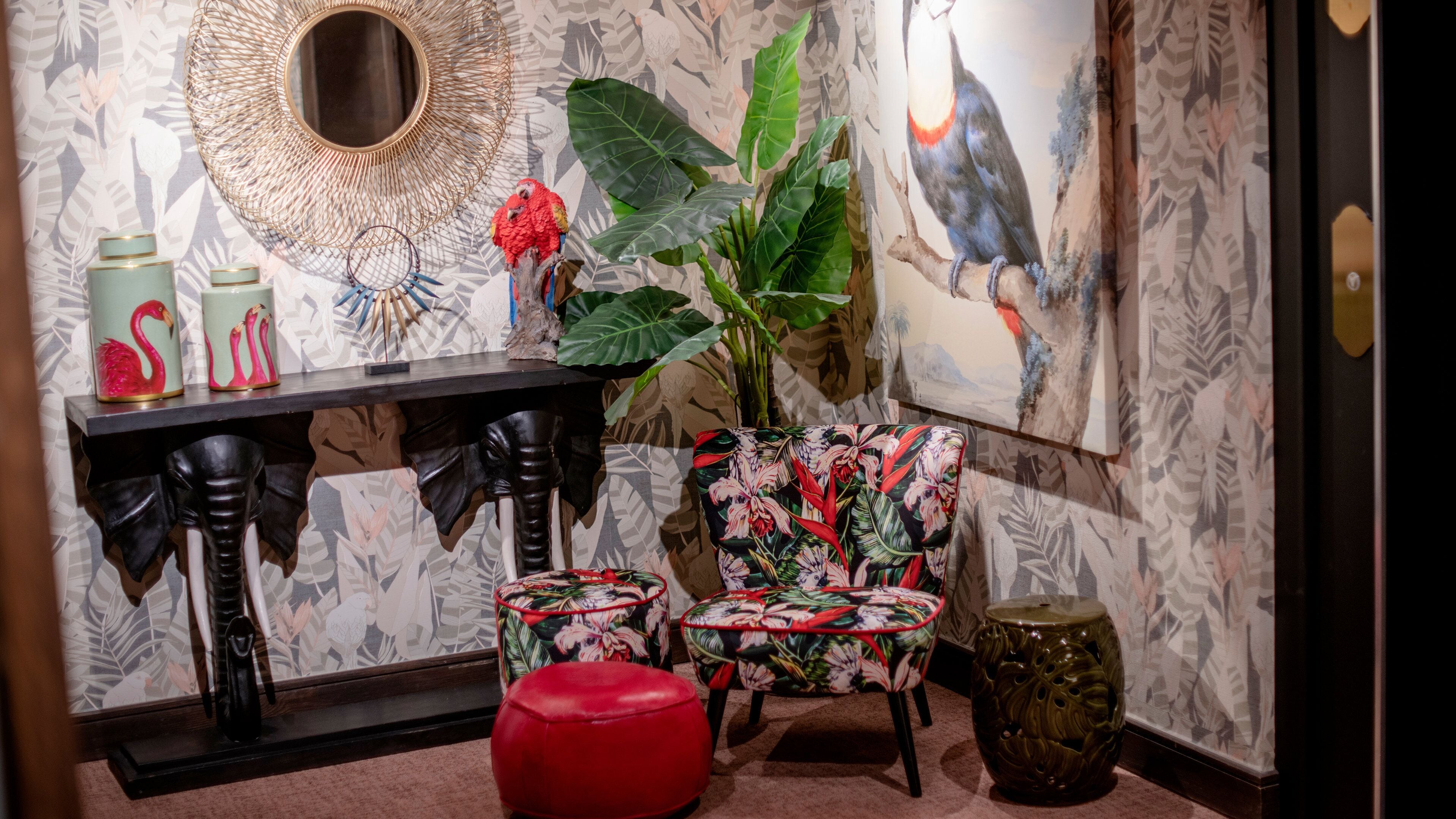 A room with a large plant, a red ottoman, a chair with a floral pattern, a console table with decorative items, and a painting on the wall.
