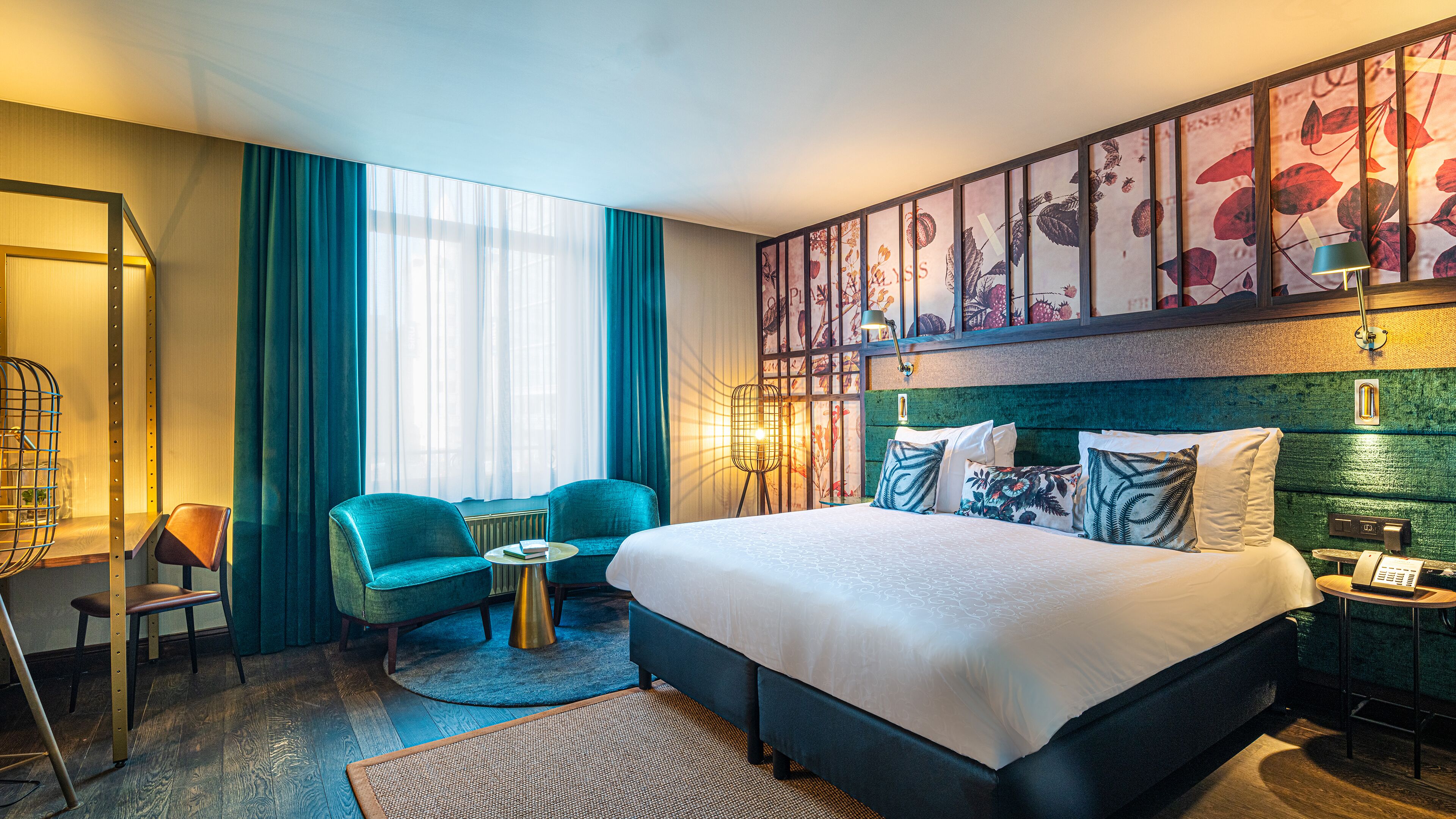 Een hotelkamer met een groot bed, twee stoelen, een klein tafeltje en een groot raam met gordijnen.