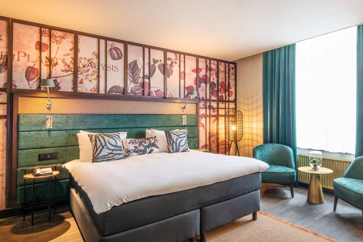 A hotel room with a large bed, two armchairs, a small table, and a wall with botanical artwork.