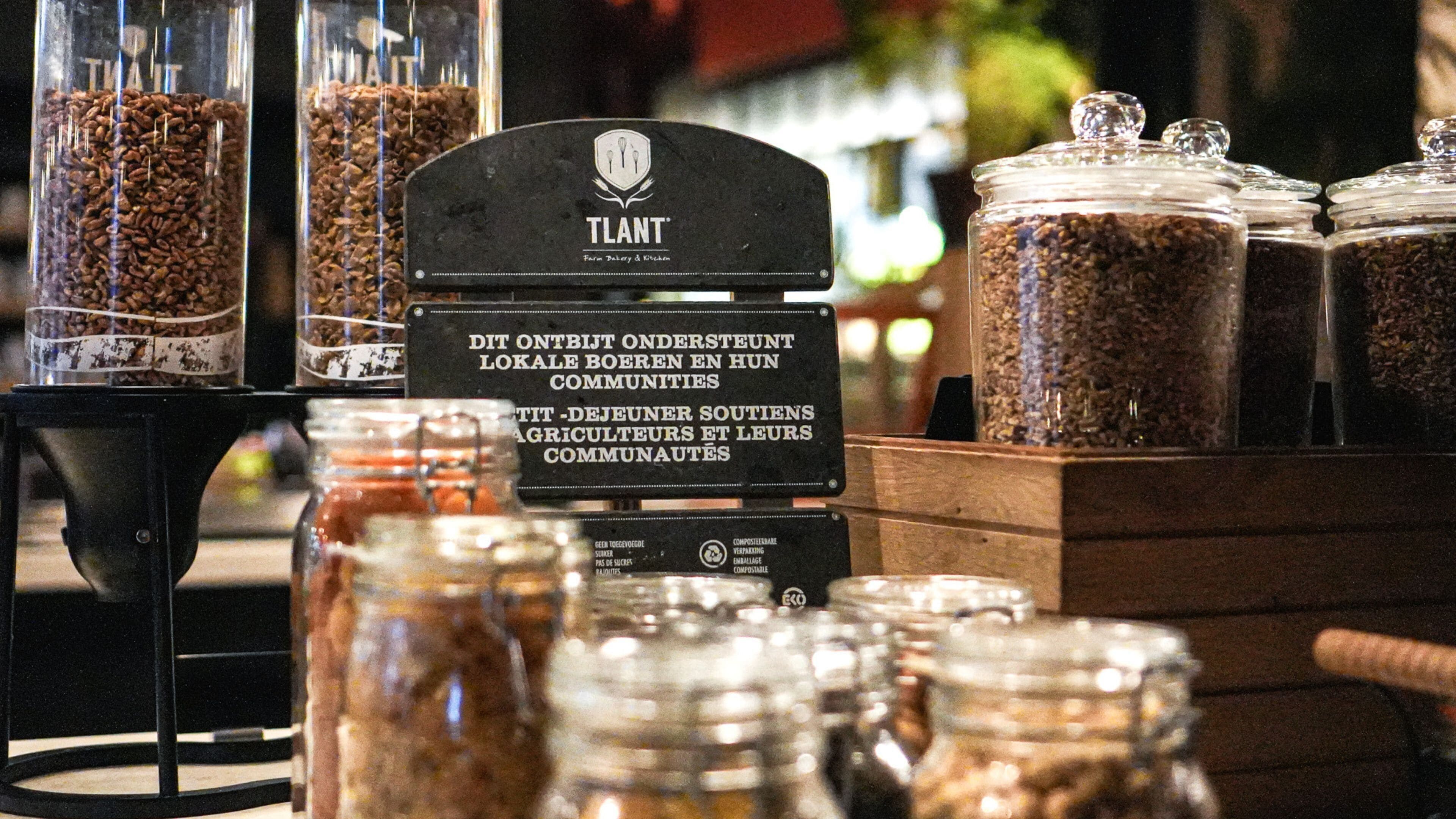 Presentación de diversos alimentos en recipientes de vidrio, incluyendo granos de café y cereales, con un cartel que indica apoyo a las comunidades locales y prácticas sostenibles.