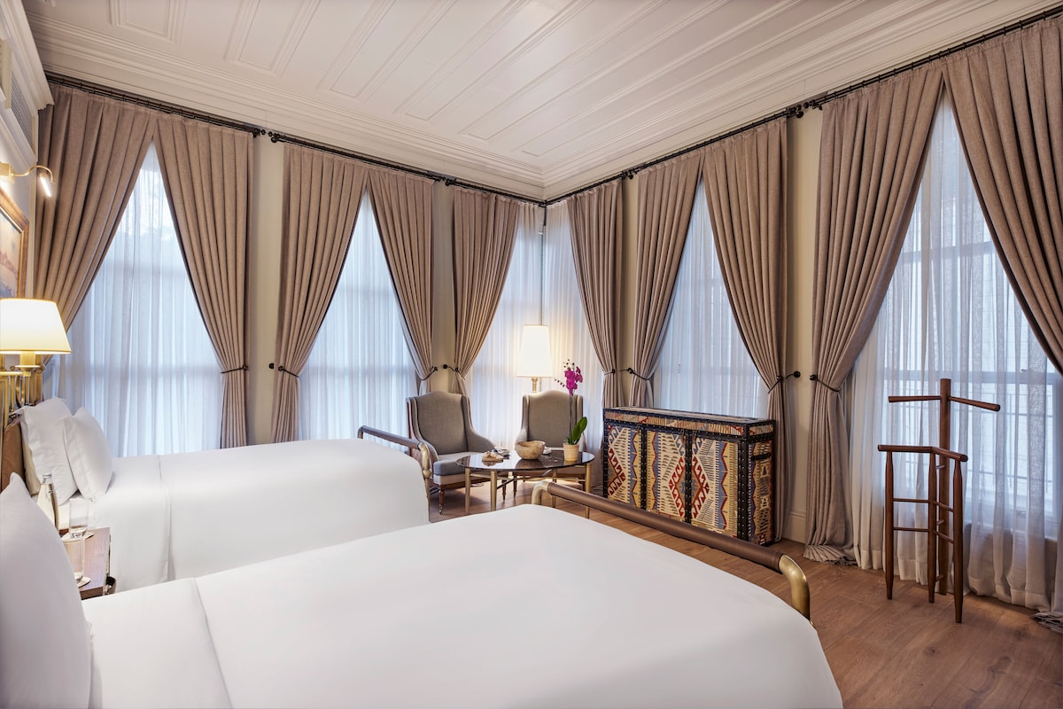 A hotel room with two beds, a sitting area with a table and chairs, a small side table with a lamp, and large windows with curtains.