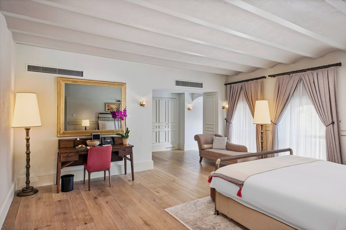 A hotel room with a wooden desk, a large bed, a sitting area, and a mirror with a gold frame.