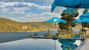 Vista clara de uma piscina com montanhas ao fundo, cercada por guarda-sóis azuis e um jardim bem cuidado.