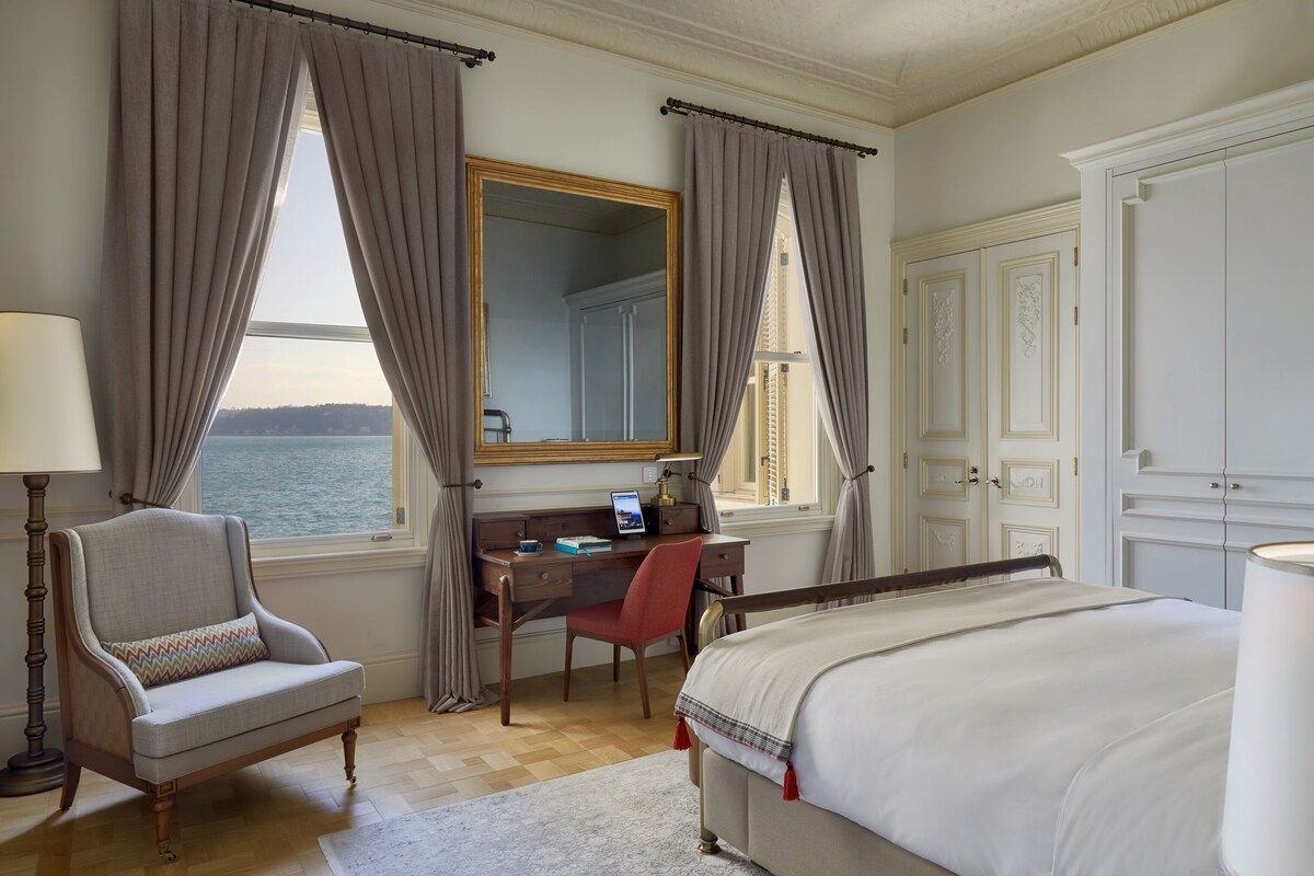 A hotel room with a bed, a desk, a chair, a lamp, and a large window with curtains.