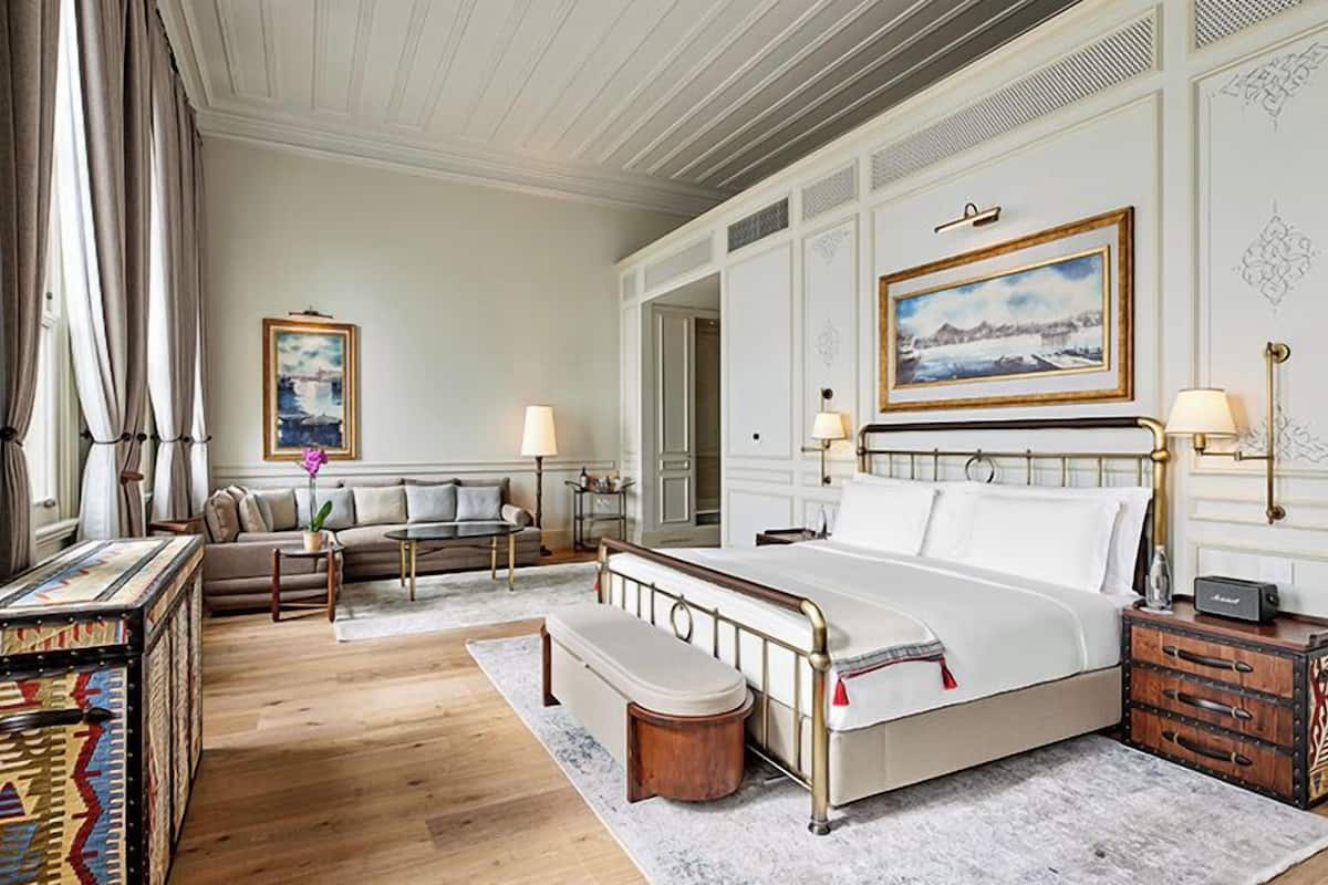 A hotel room with a bed, bedside tables, a bench, a dresser, a sofa, a coffee table, and a painting on the wall.