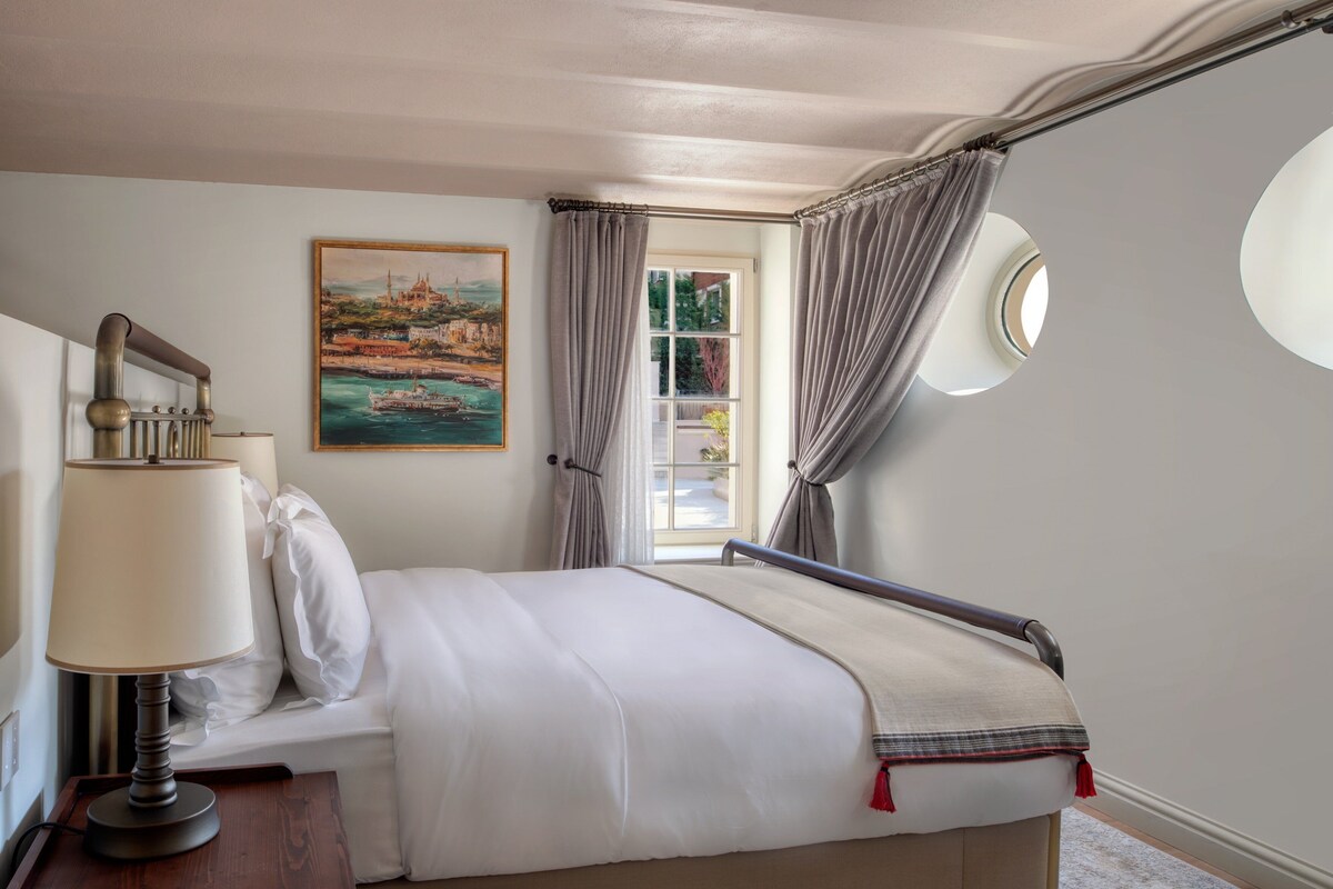 A neatly made bed with a headboard, a bedside lamp, a painting of a coastal scene, and a window with curtains.