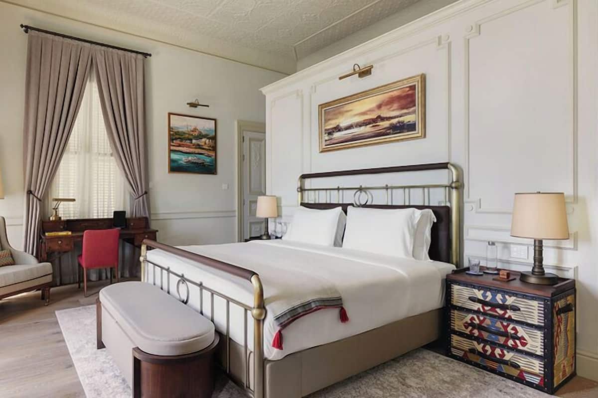 A hotel room with a bed, a chair, a desk, a lamp, and a painting on the wall.