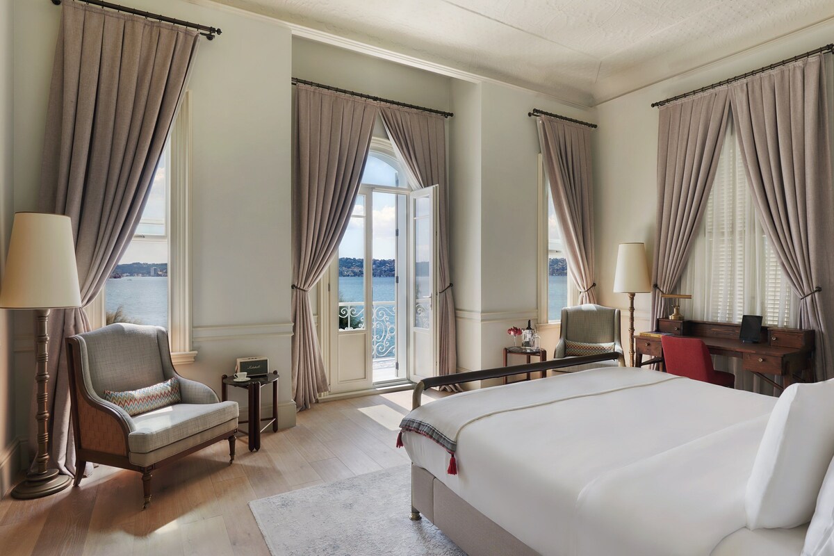 A hotel room with a large bed, a chair, a desk, and a view of the sea.